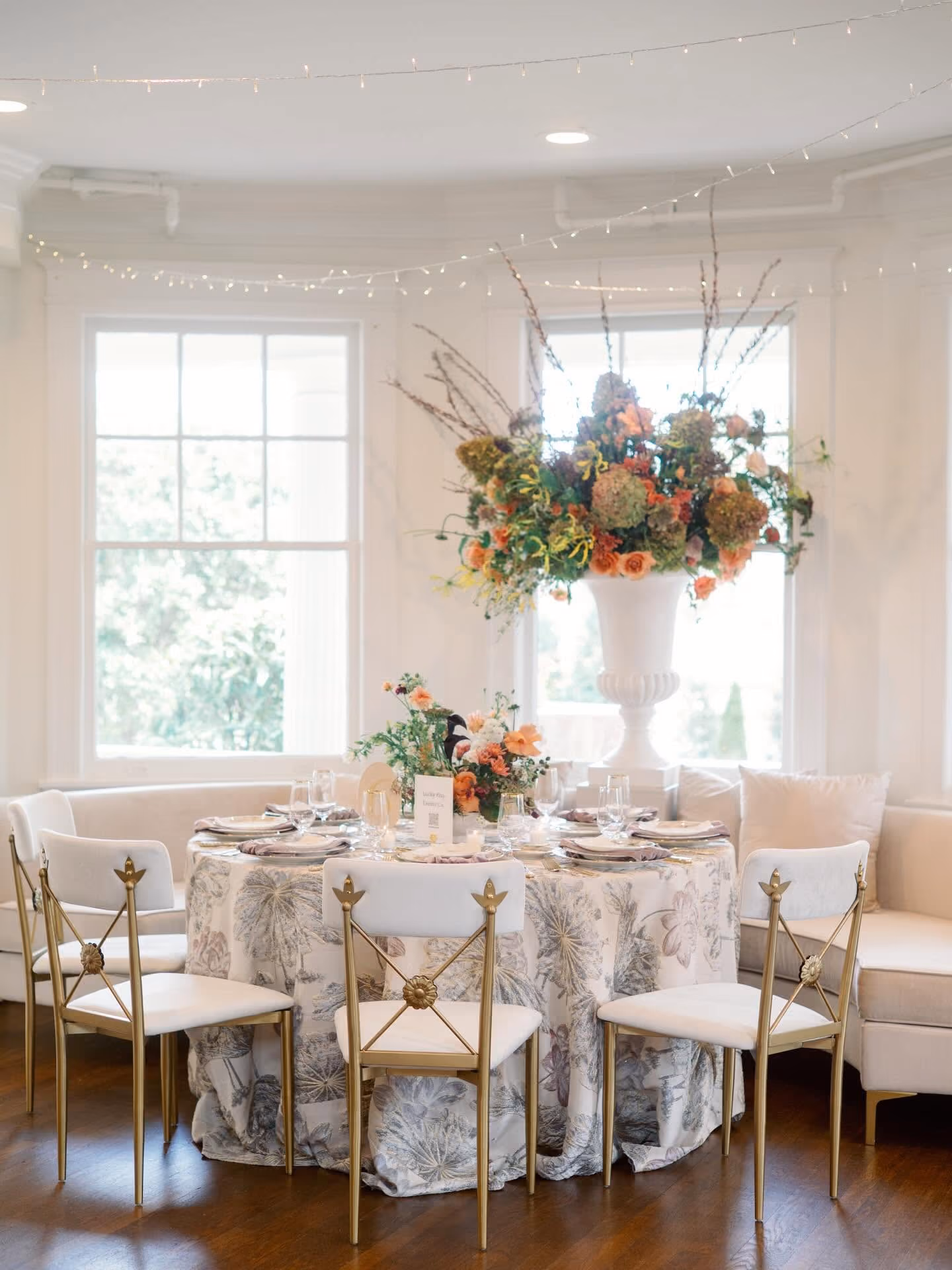 Elegant round table with floral tablecloth, set with glassware and plates, surrounded by gold chairs and floral centerpieces.