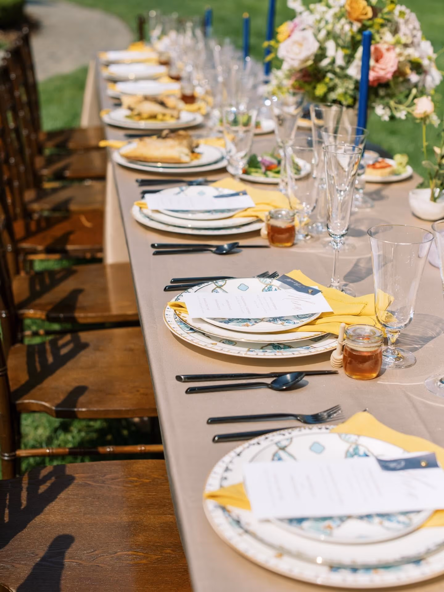 Outdoor dining table set with patterned plates, yellow napkins, glassware, and honey jars under sunlight.