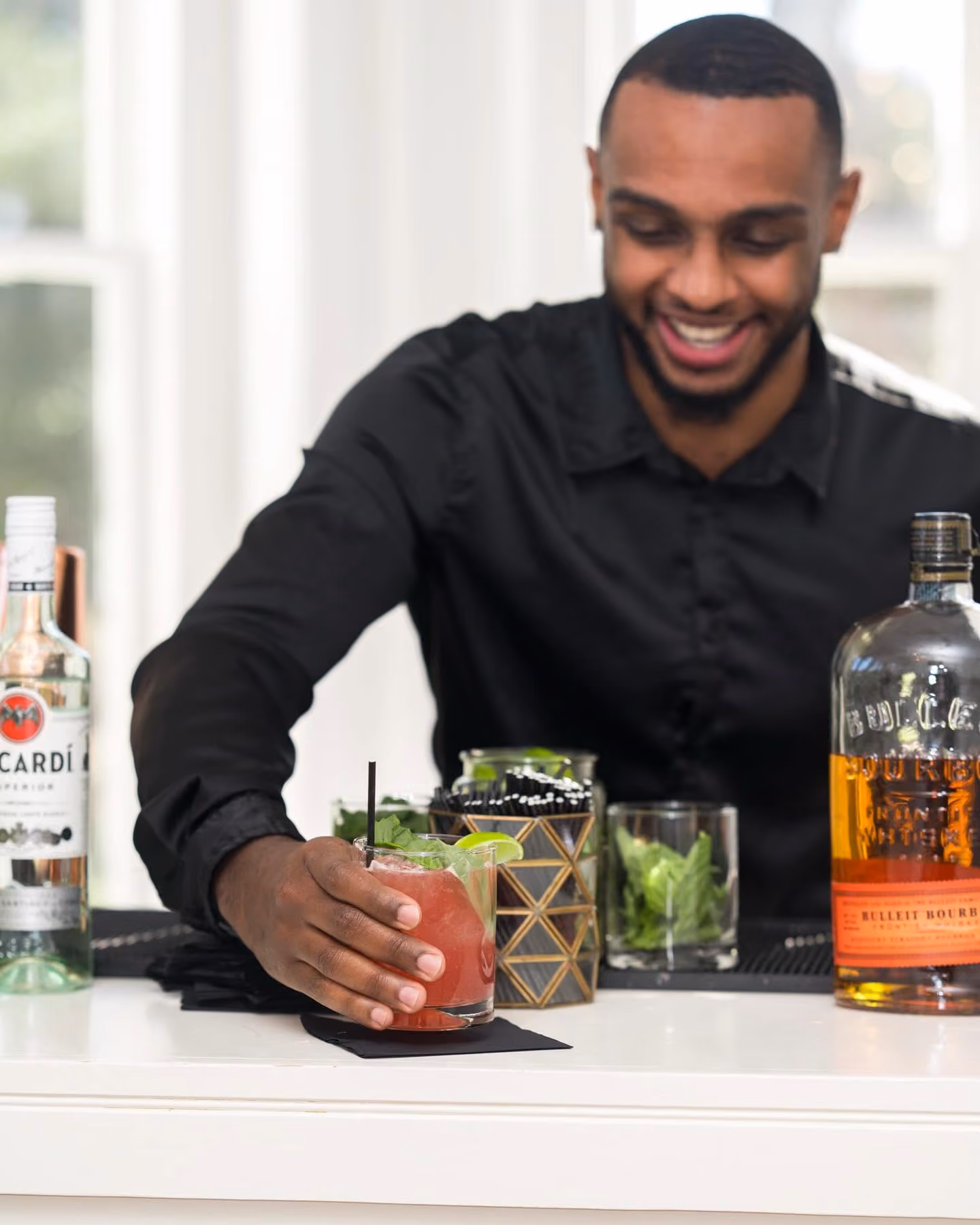Smiling bartender in black shirt holding a red cocktail garnished with lime and mint at a bar.