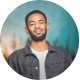 Smiling man with beard wearing a dark shirt and jacket against a blue blurred background.