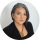 Woman with shoulder-length gray hair wearing a black blazer and gold necklace against a gray background.