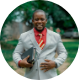 Smiling man in light gray suit holding a tablet, standing outdoors with green trees in background.