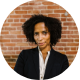 Smiling person with curly hair wearing a black blazer and white shirt against a brick wall.