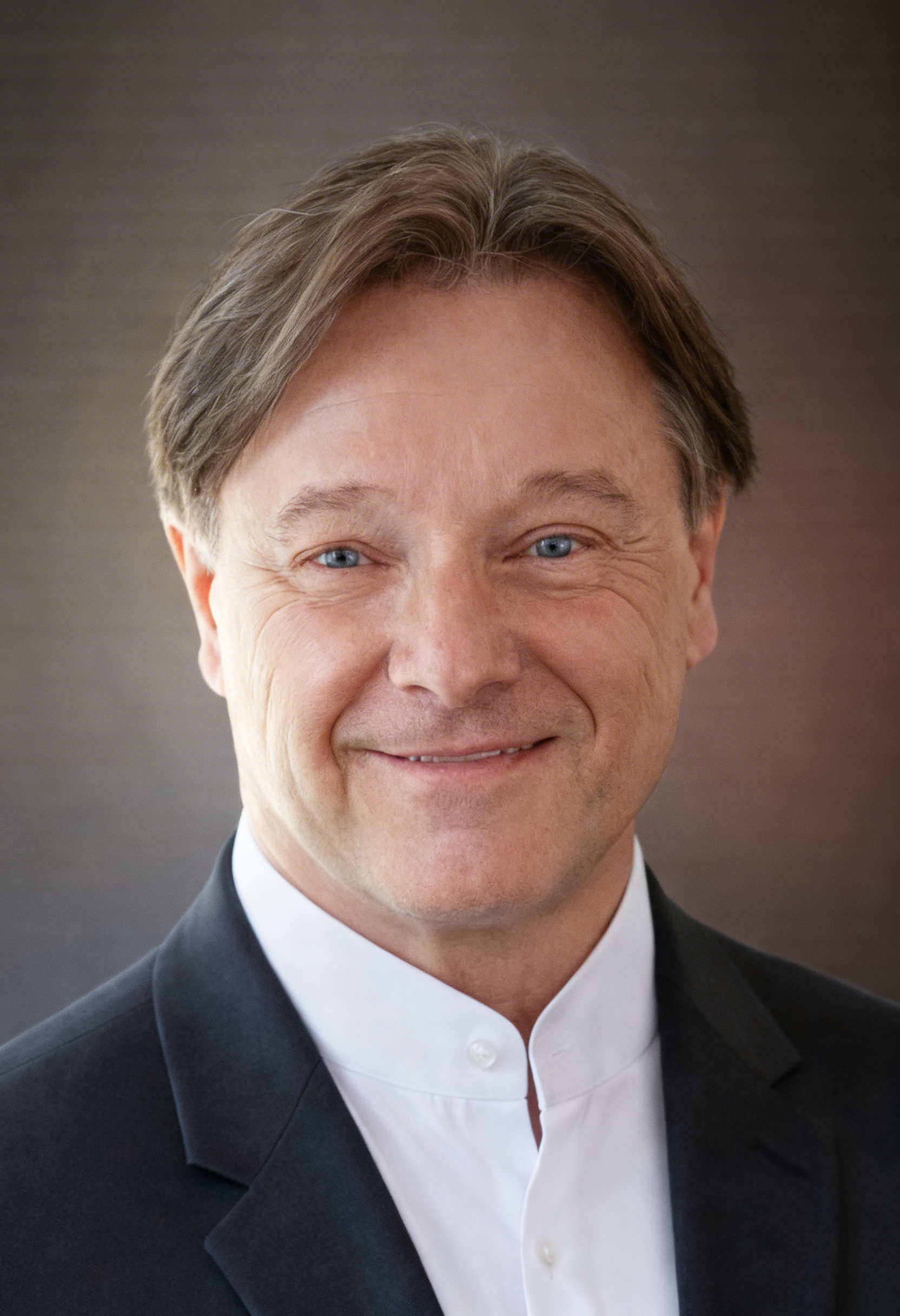 Middle-aged man with light brown hair and blue eyes wearing a white shirt and dark blazer, smiling at the camera.