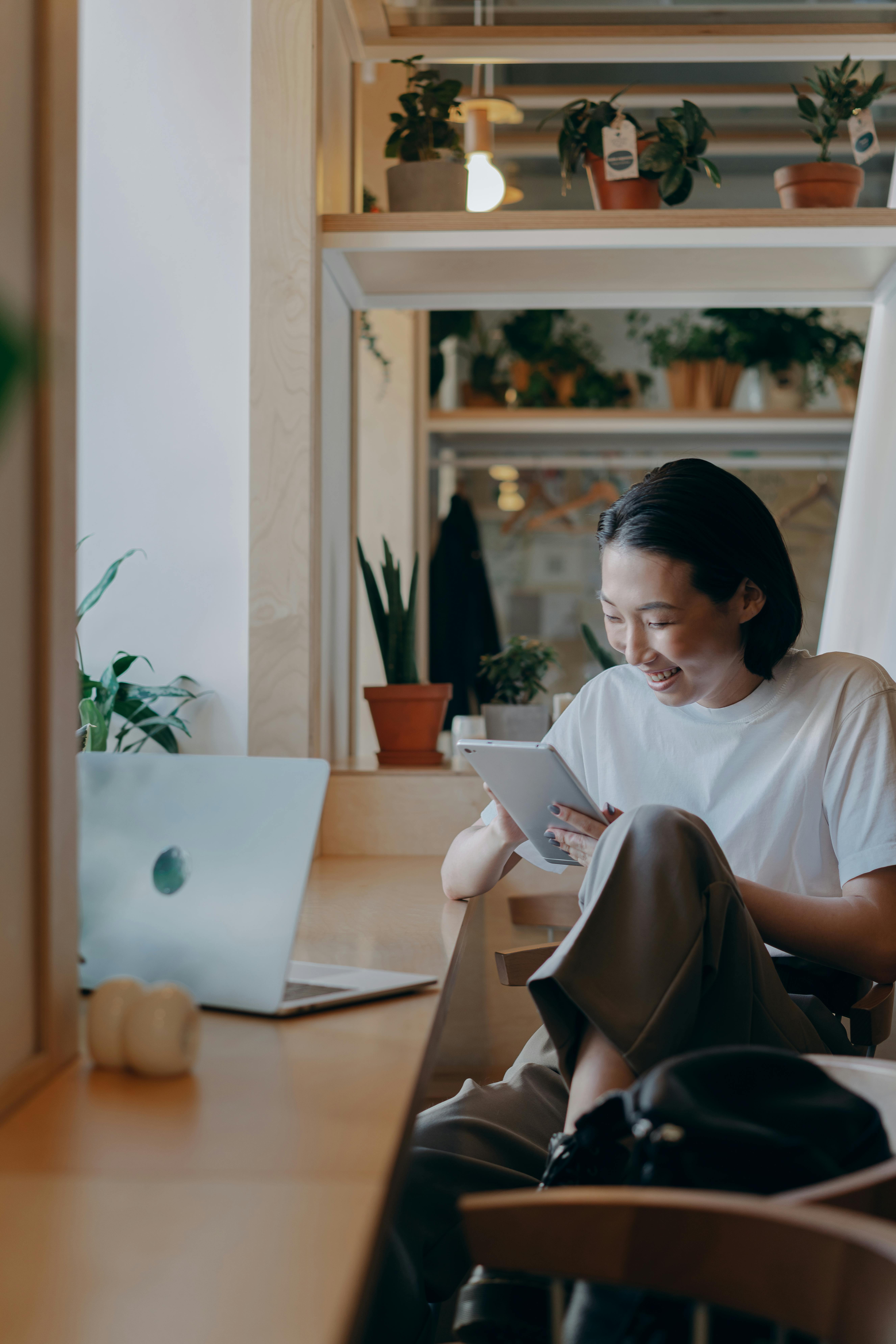 Person smiling while sitting at a wooden desk holding a tablet, with a laptop and potted plants nearby in a well-lit modern room.