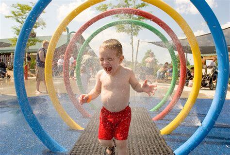 Young boy in red shorts playing and running through colorful water spray rings at a splash pad on a sunny day.