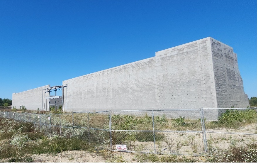 Large unfinished concrete building behind a chain-link fence with dry grass and clear blue sky.