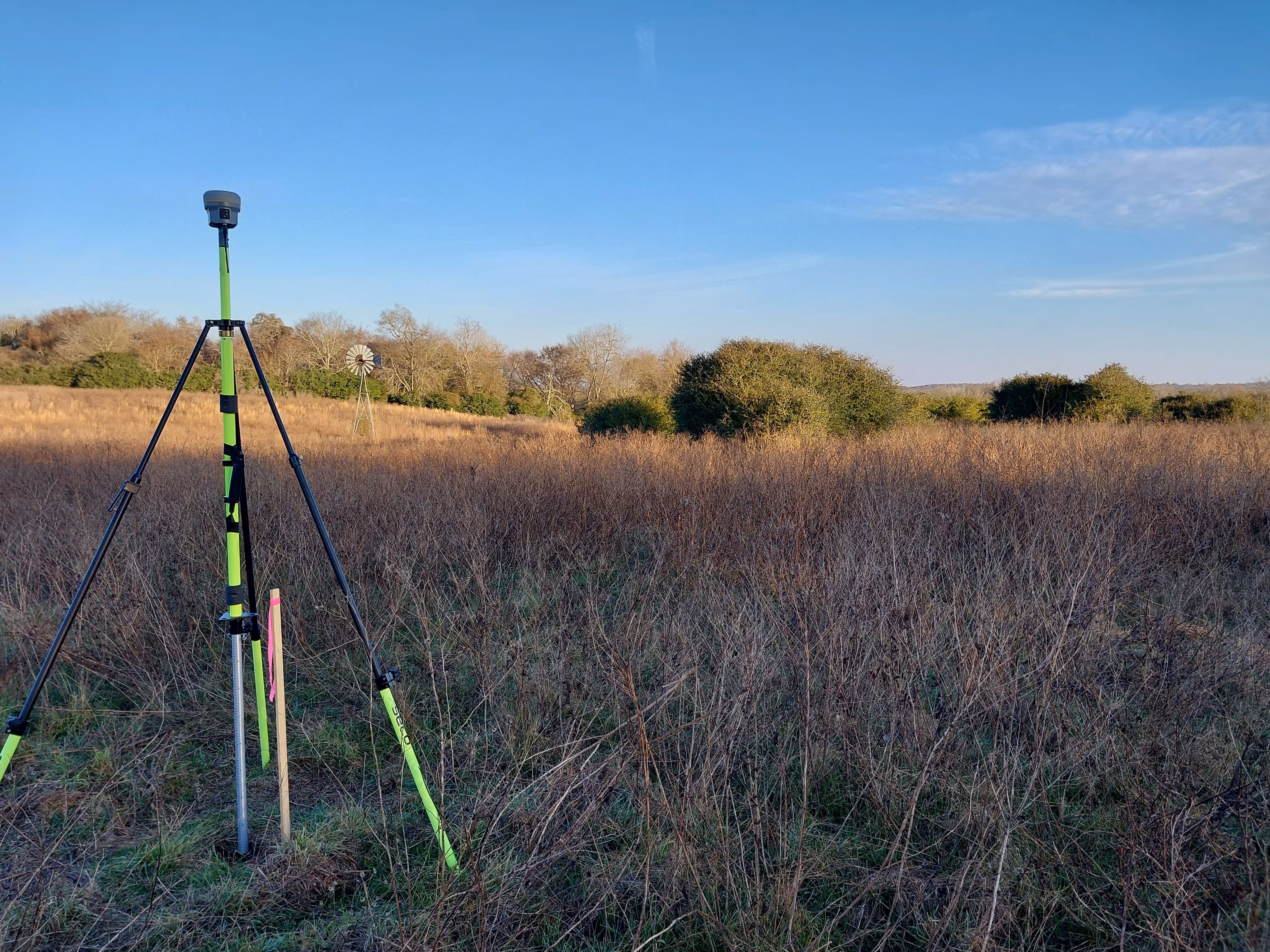 Trimble R10 GNSS receiver collecting raw static data over a known survey control point in an open field for aerial mapping.
