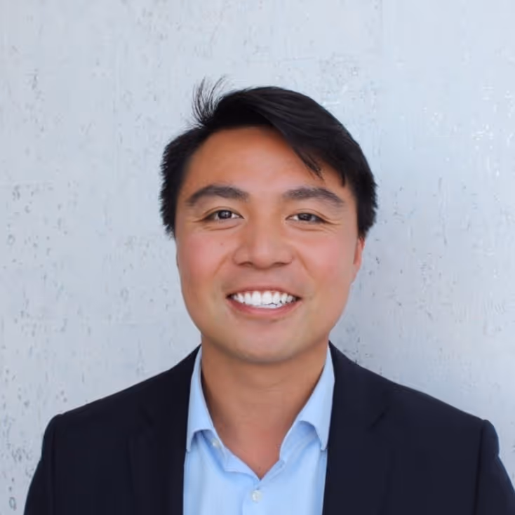 A person smiling warmly at the camera against a textured white background. They wear a dark blazer and light blue shirt. Previously with Wunderkind, placed with Unify.