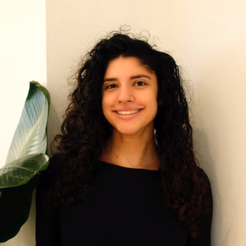 Smiling, professional woman with curly hair stands beside a leafy plant against a light wall. Previously with Larson International Group, placed with Warp