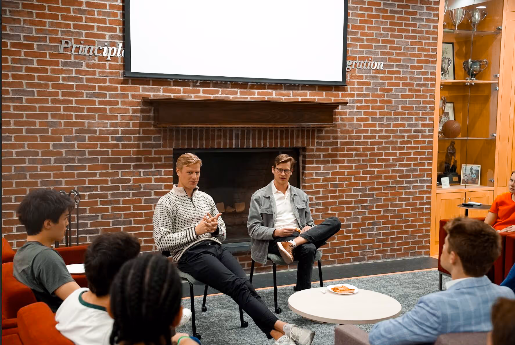Two men sit in front of a group of candidates in a cozy room at Harvard University with a brick wall and fireplace. One speaks animatedly, educating students about sales roles.