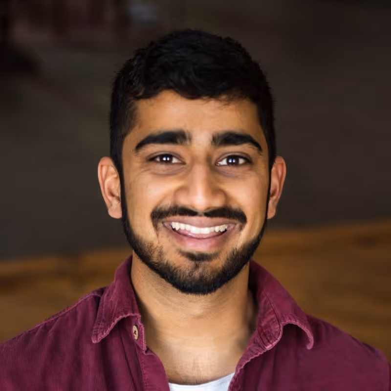 A professional man in a casual red button down shirt smiling in front of a blurred brown background.