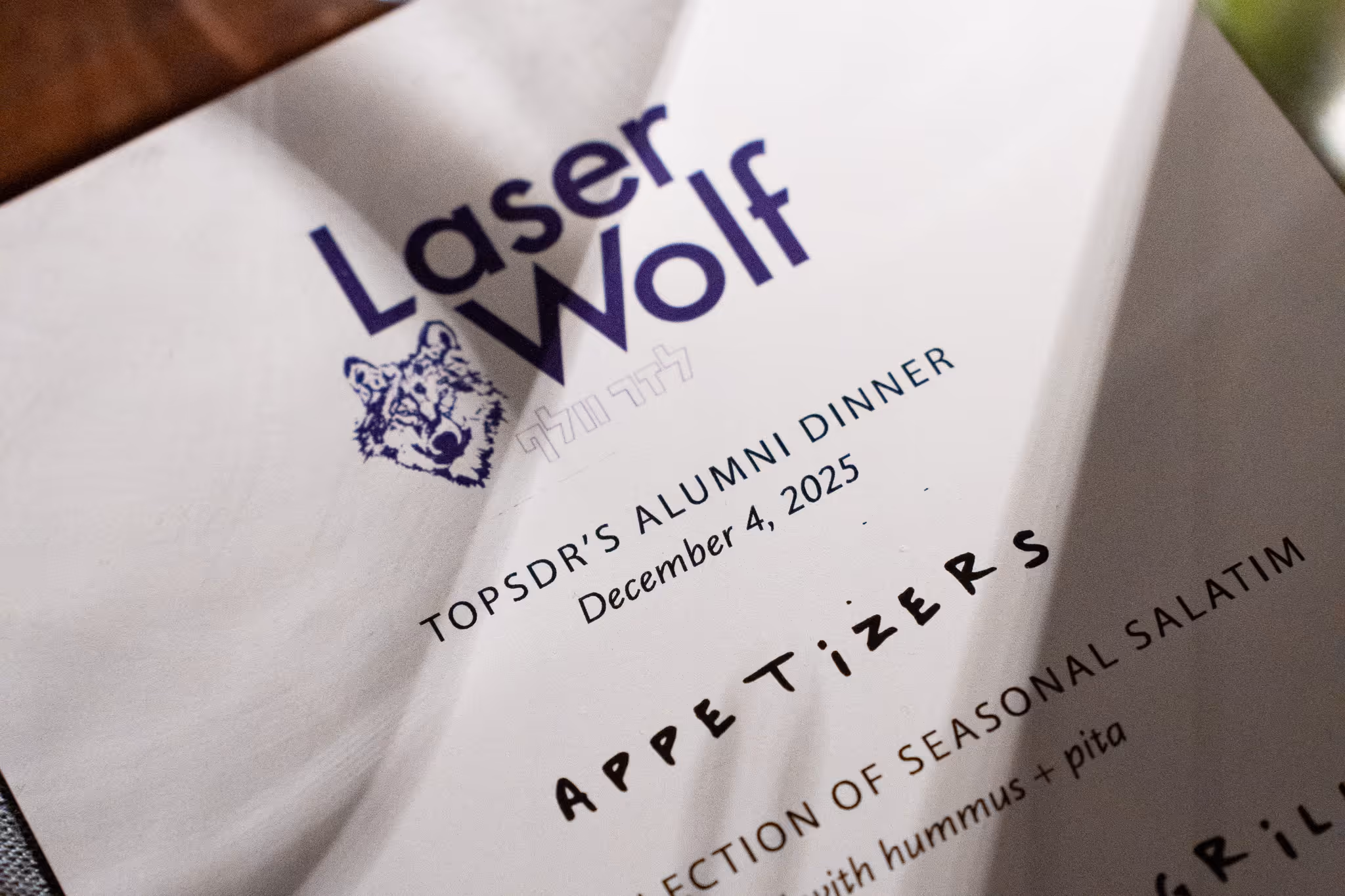 Close-up of a menu titled "Laser Wolf" featuring a wolf logo. It notes "TOPSDR's Alumni Dinner" on December 4, 2025, with "Appetizers" section below.