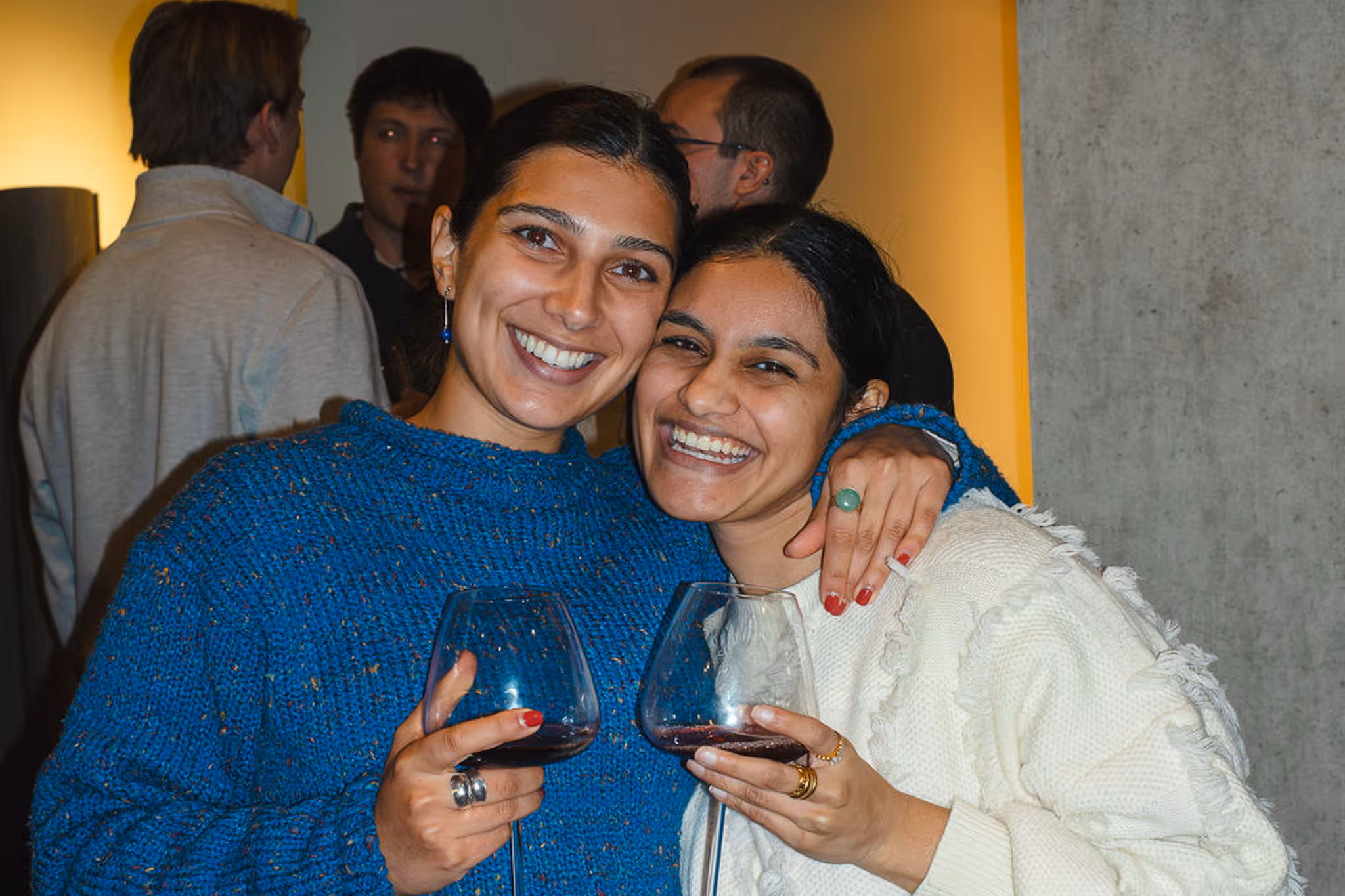 Two people smiling and holding glasses of red wine, hugging at a gathering for young professionals. They wear cozy sweaters, creating a warm and joyful atmosphere.
