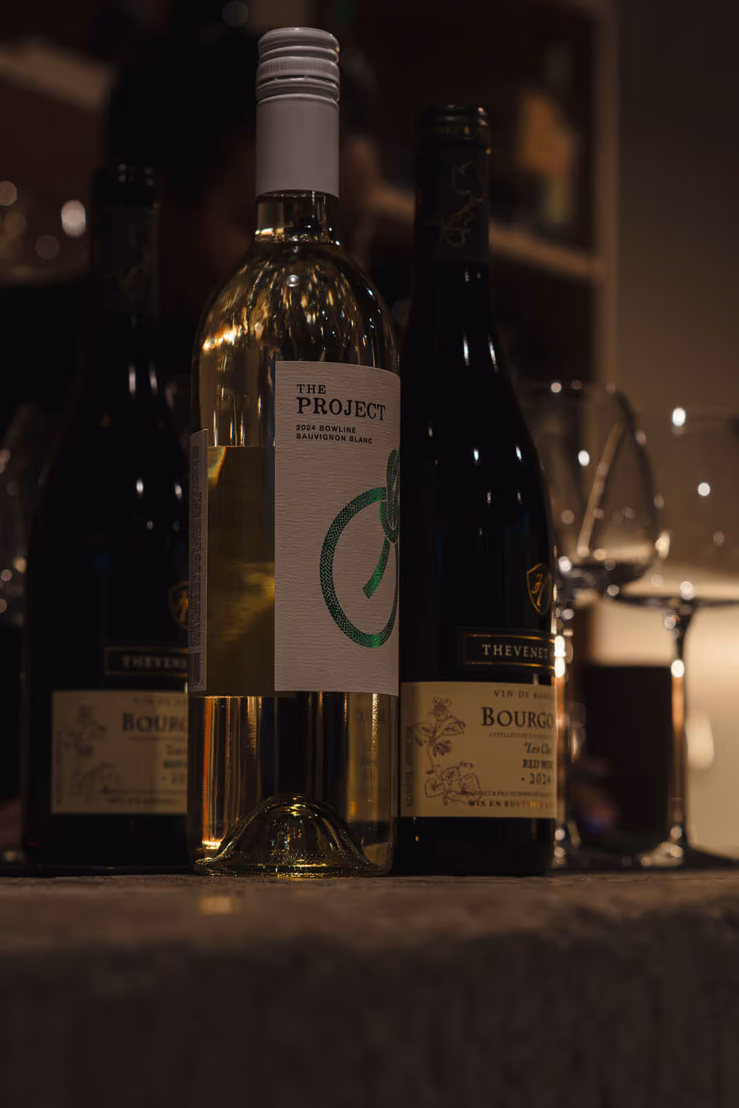 Three bottles of wine on a table with a focus on a white wine bottle labeled The Project 2024 Bowline Sauvignon Blanc, with two wine glasses in the background.
