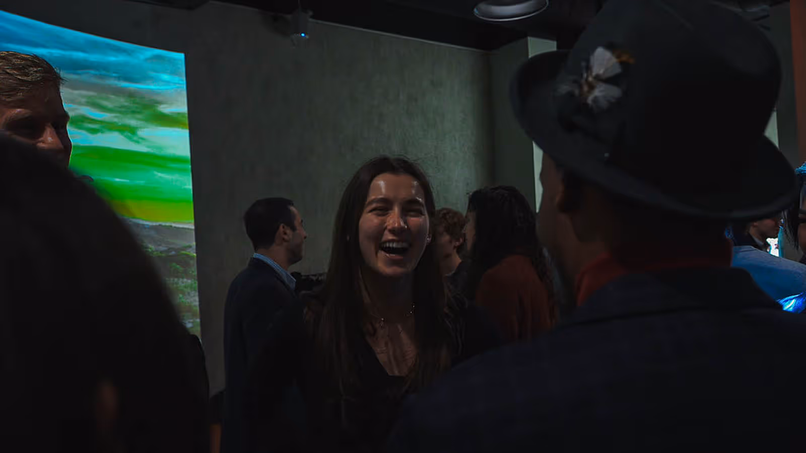 A woman laughing and conversing with a group of people in a dimly lit room with a colorful projection on the wall.