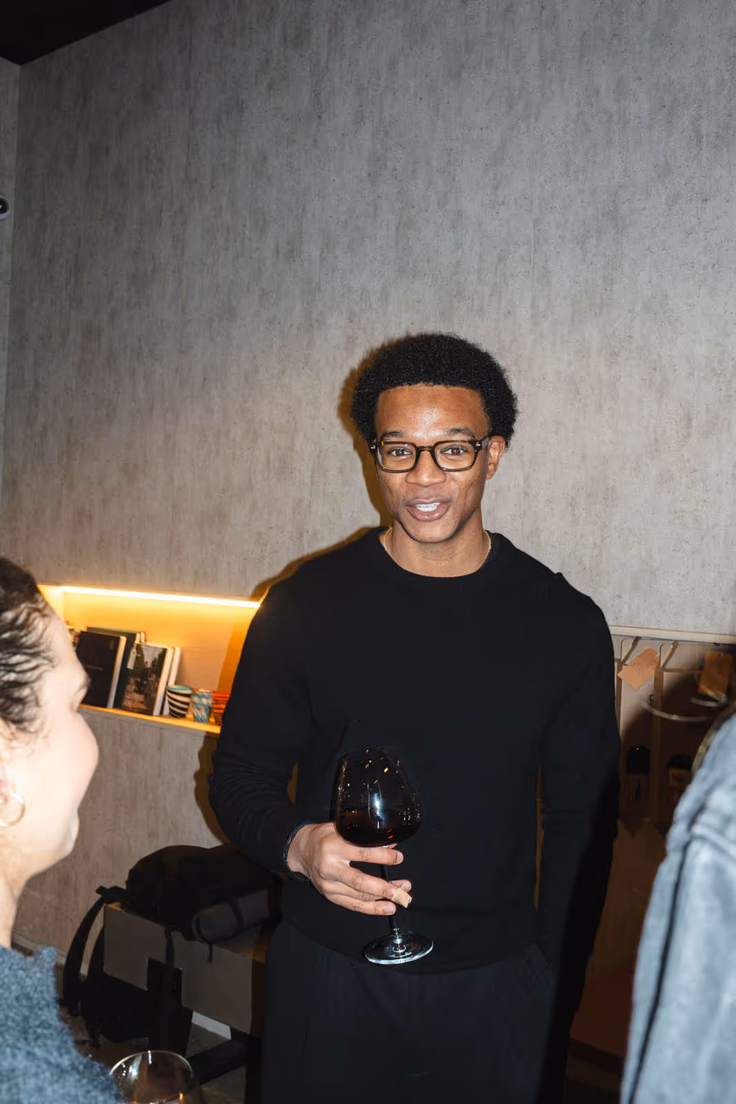 Young man wearing glasses and a black sweater holding a glass of red wine, talking with others indoors.