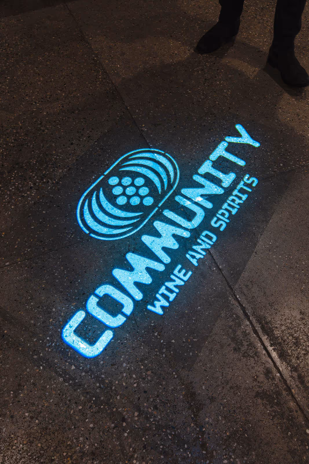 Glowing blue projection on pavement displaying 'COMMUNITY WINE AND SPIRITS' with a circular logo above the text.