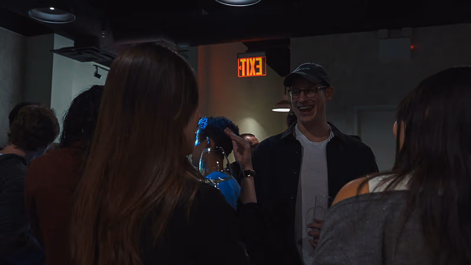 People socializing indoors under an illuminated EXIT sign, one man in a cap smiling and holding a drink.