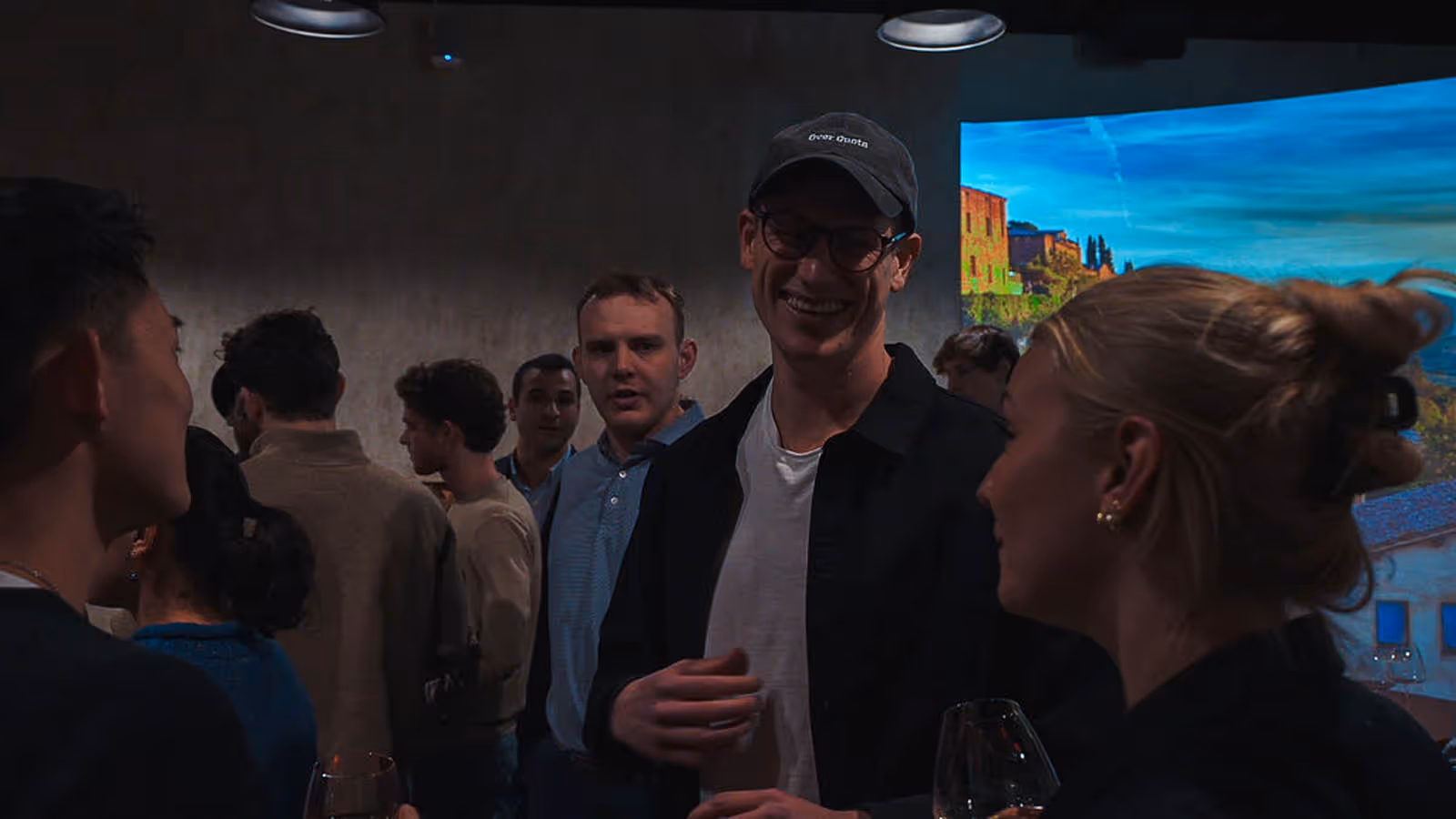 Group of people socializing indoors, one man with glasses and a cap smiling in the center, others engaged in conversation around him.