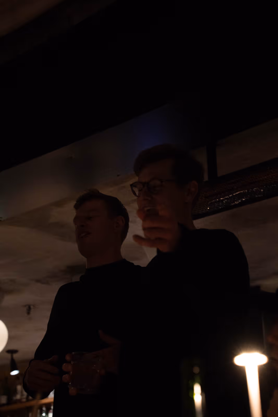 Two men standing and talking in a dimly lit indoor setting, one holding a glass.