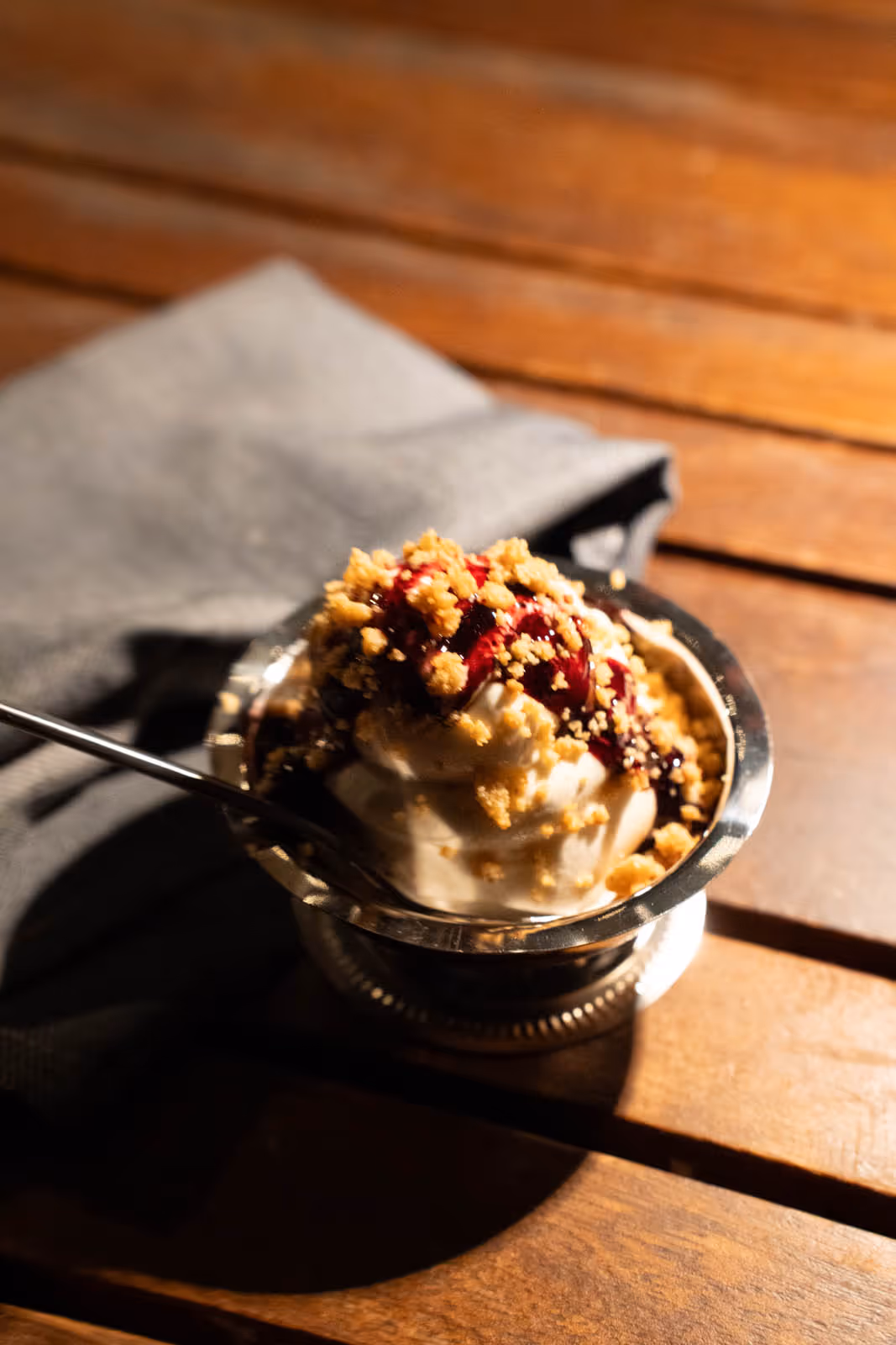 Metal bowl of vanilla ice cream topped with berry sauce and crunchy crumbs on a wooden table with a spoon.
