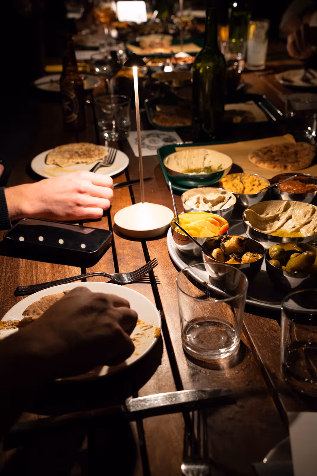 Wooden table with various dips, pickles, and flatbreads served in metal bowls, with two people eating in dim lighting.