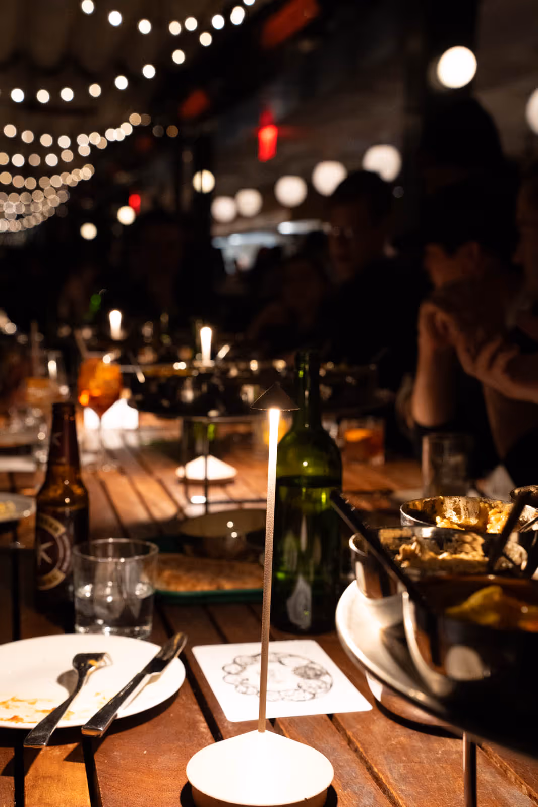 Dimly lit wooden table set with dishes, glasses, a bottle, and a small tall candle at a gathering with string lights overhead.