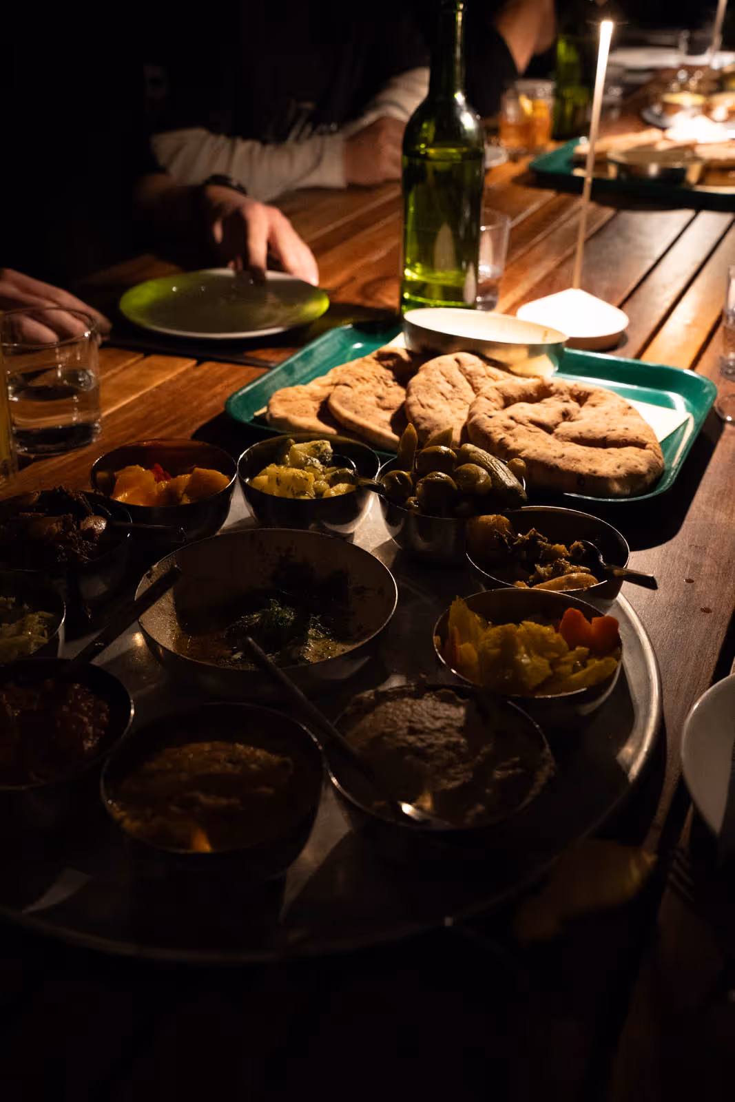 Tray with pita bread, various small bowls of dips and pickled vegetables on a wooden table with dim lighting.