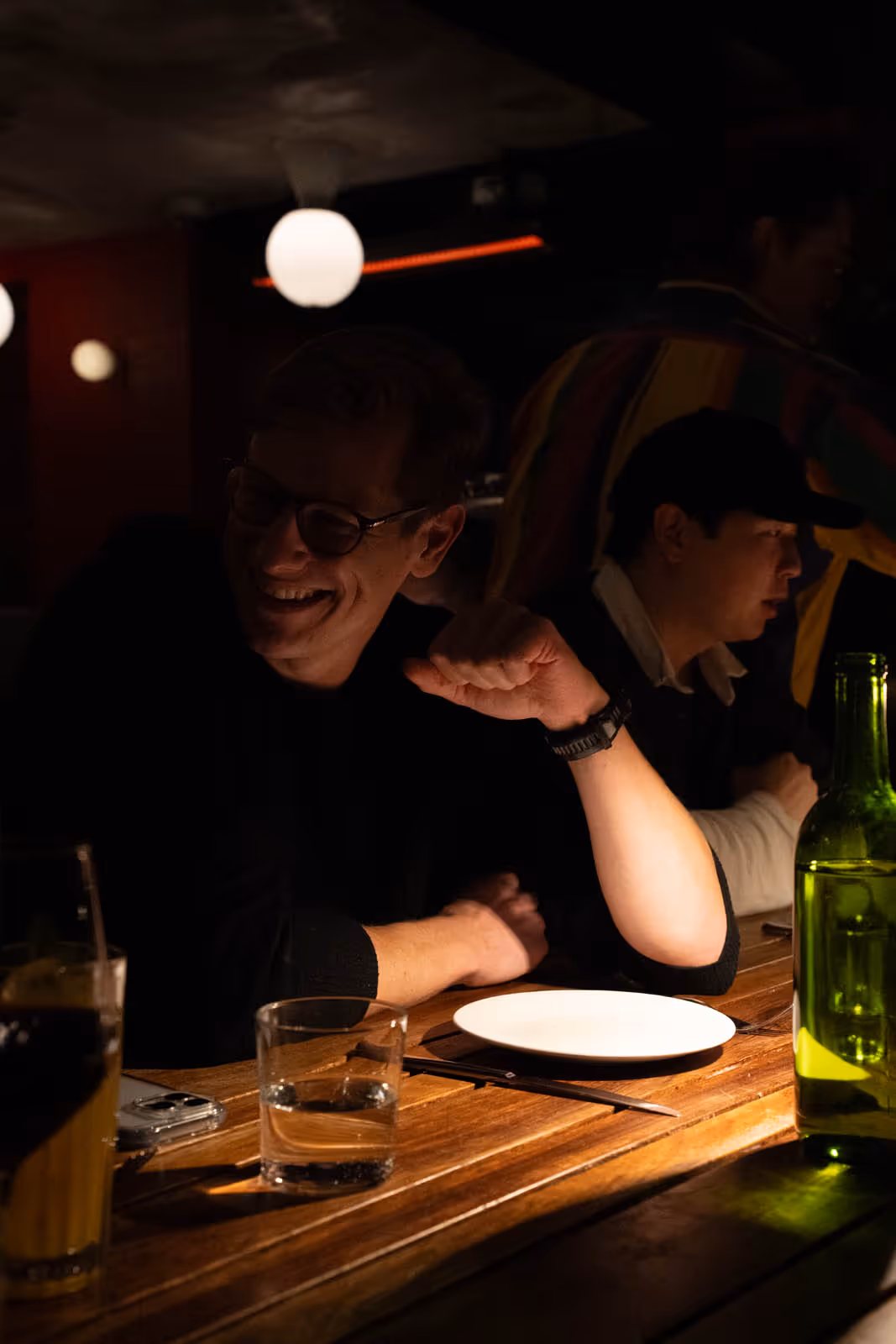 Two men sitting at a wooden table in a dimly lit bar, one smiling with glasses and the other wearing a black cap.