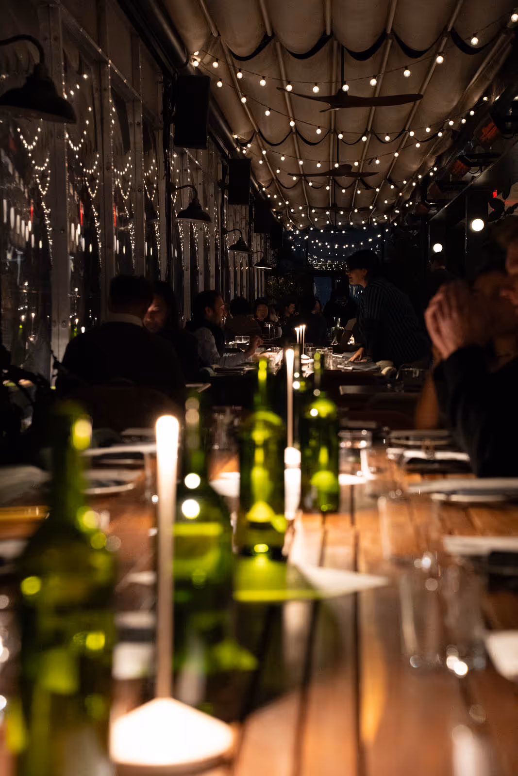 Dimly lit restaurant interior with string lights overhead and green glass bottles on a wooden table.