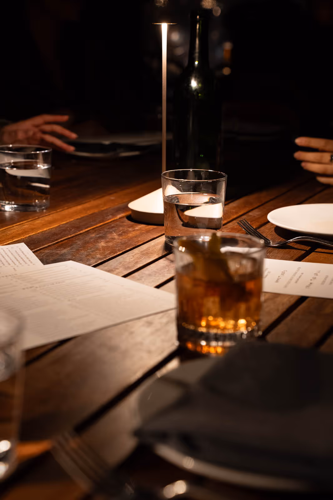 Dimly lit wooden table with two glasses of water, a glass of amber drink with lemon, menus, a small lamp, a bottle, and a fork with a white plate.