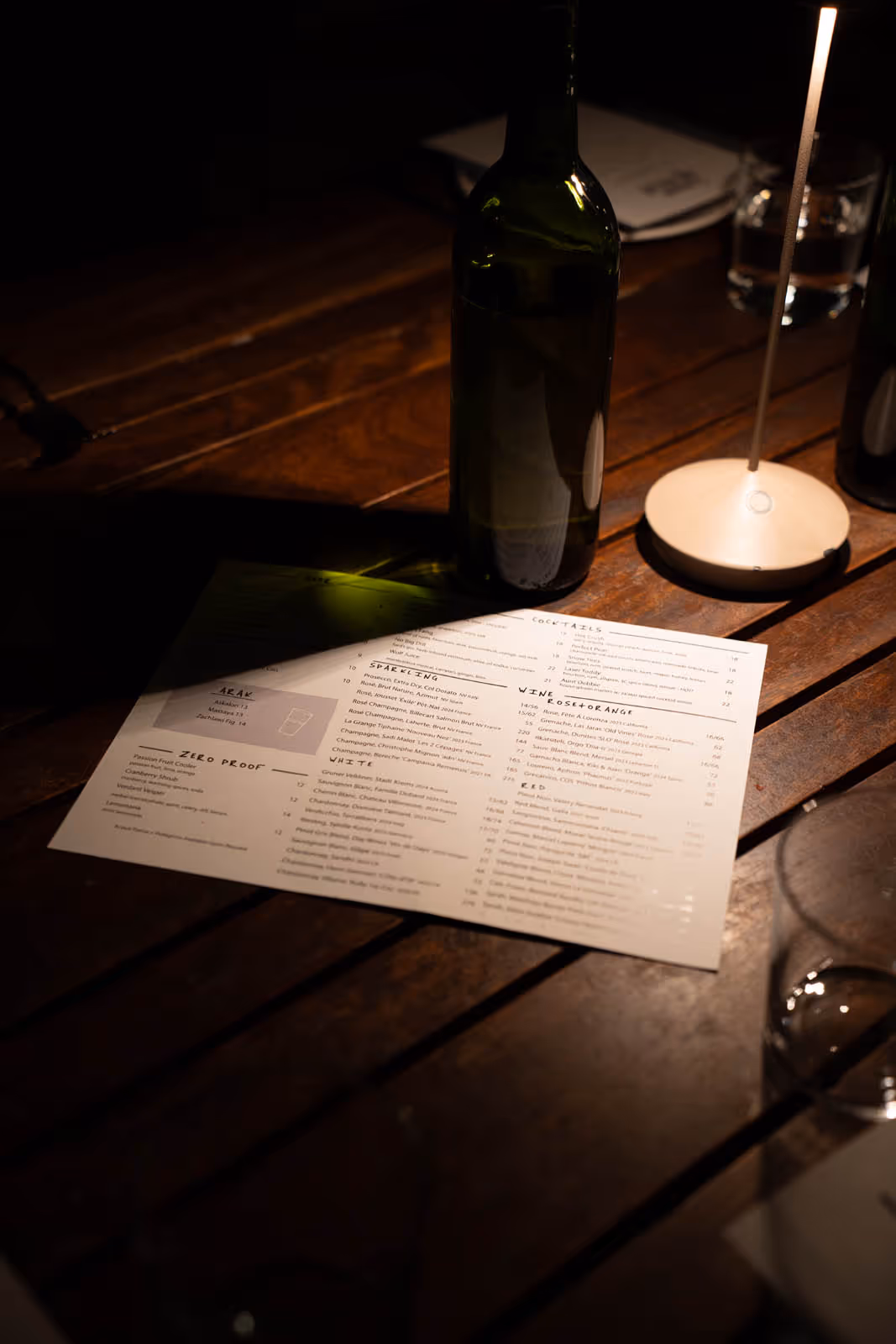 Dimly lit wooden table with a wine bottle, a menu, and a small lamp casting light on the menu.