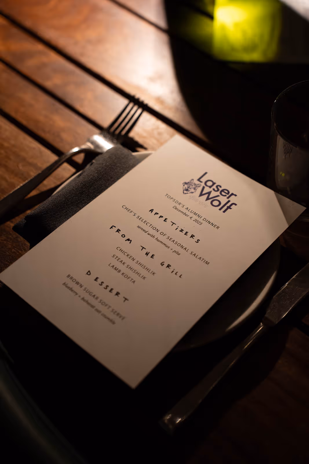 A restaurant menu titled 'Laser Wolf' for Topsor's Alumni Dinner on December 4, 2025, listing appetizers, grilled dishes, and dessert on a plate with fork, knife, and napkin on wooden table.