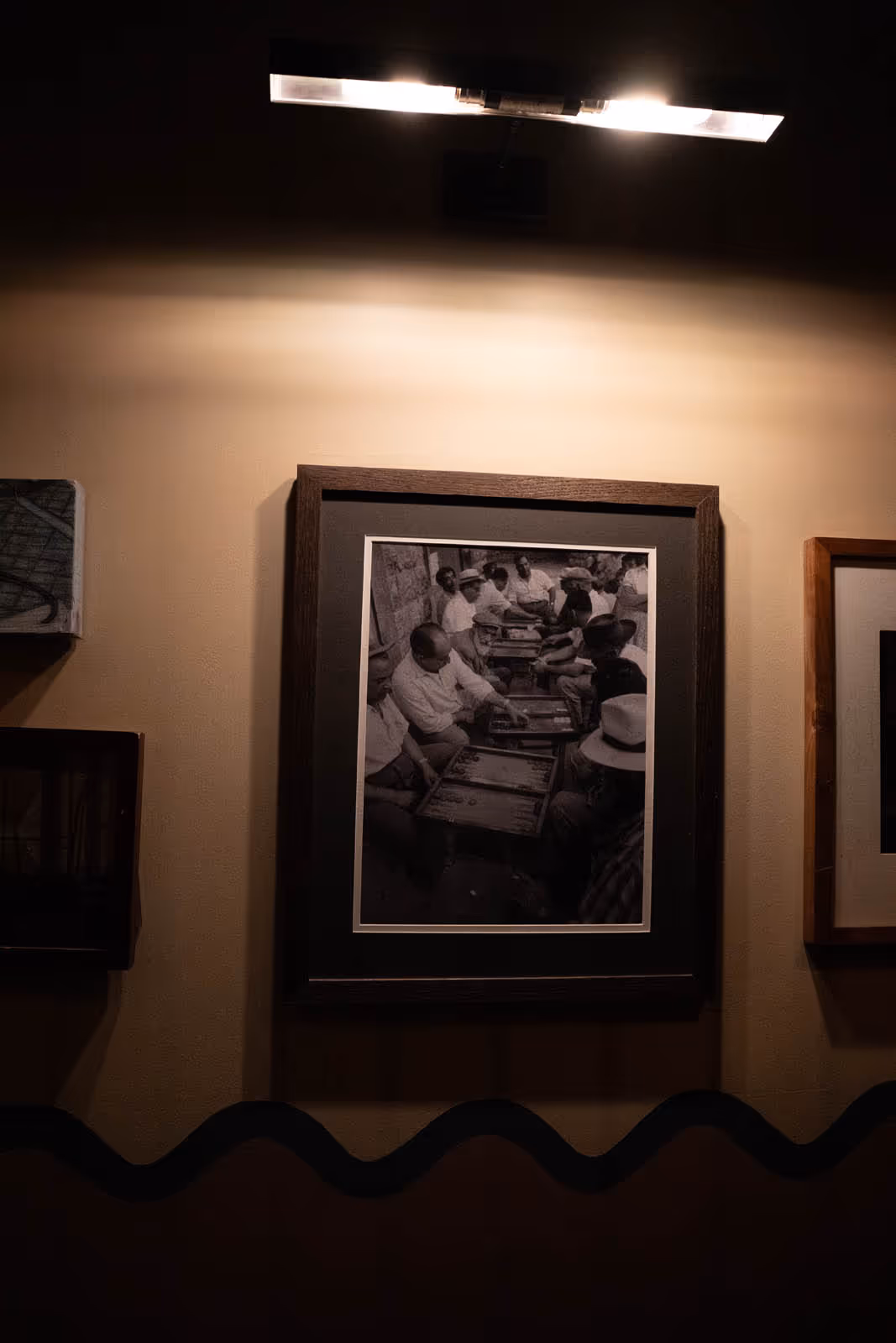 Framed black-and-white photograph of a group of people playing backgammon, mounted on a wall under a light.