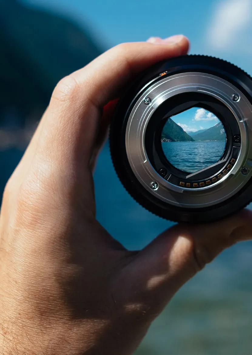 Hand holding a camera lens overlooking a lake surrounded by mountains visible through the lens.