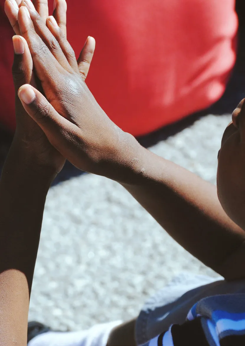 Mãos de criança negra juntas em posição de oração com fundo desfocado em vermelho e cinza.