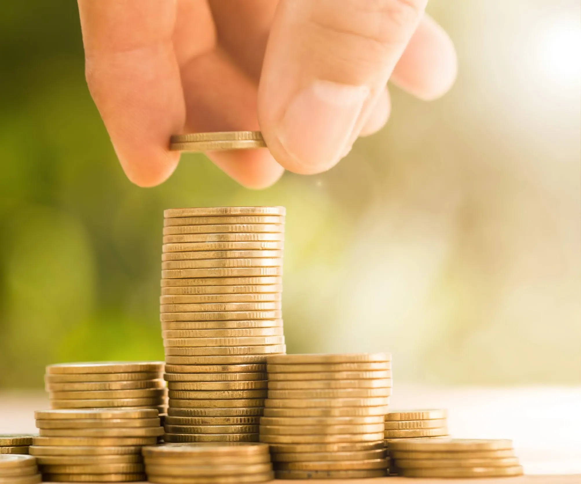 Hand placing a coin in a tall pile of gold coins with several other stacks of coins surrounding it, on a clear surface.