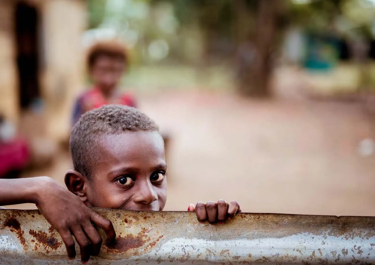 Black boy holding a rusty sheet of metal and looking directly at the camera, with another child blurred in the background.