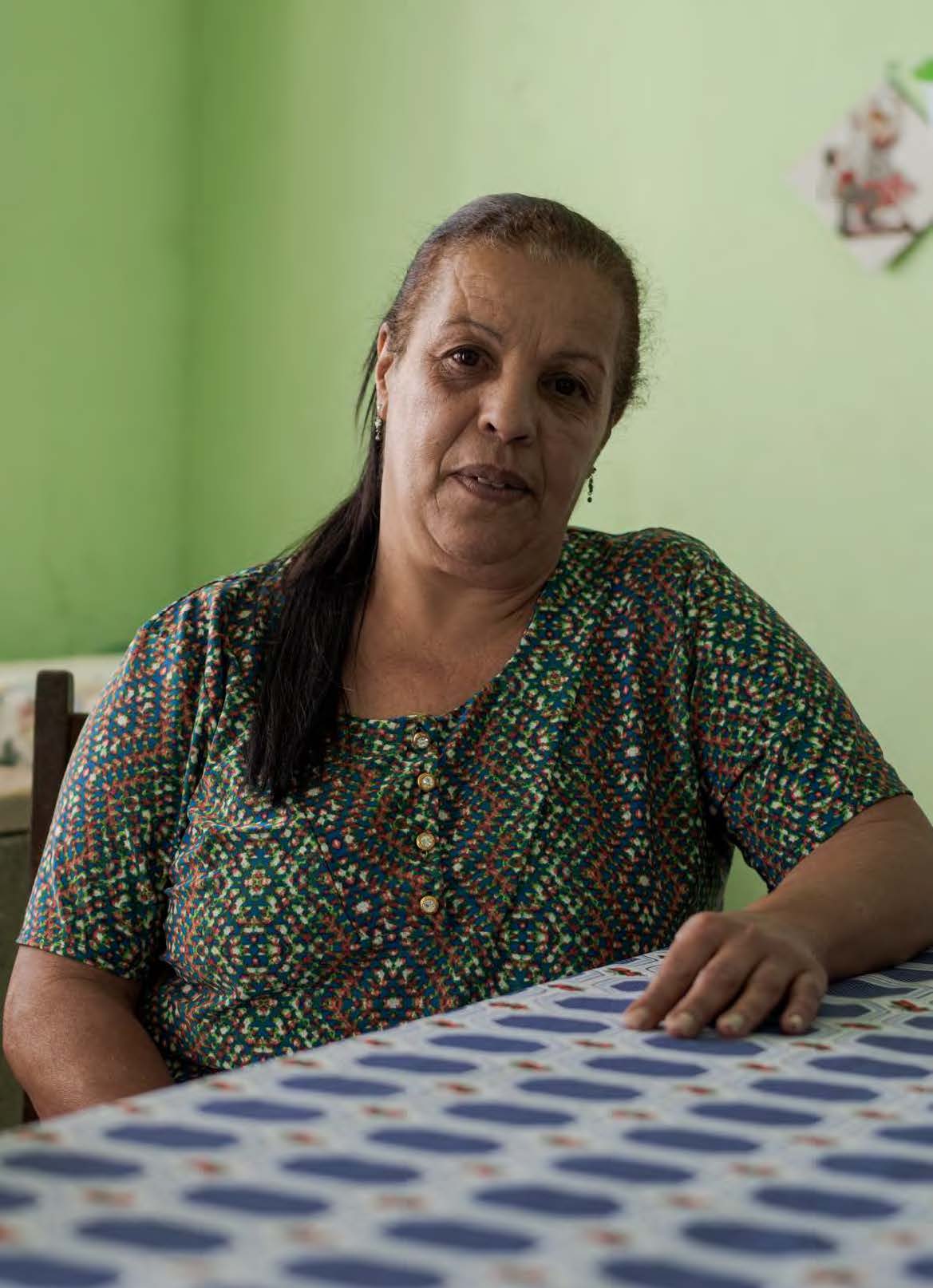 Foto da Rosilda sentada à mesa com toalha azul e branca em uma parede verde ao fundo.
