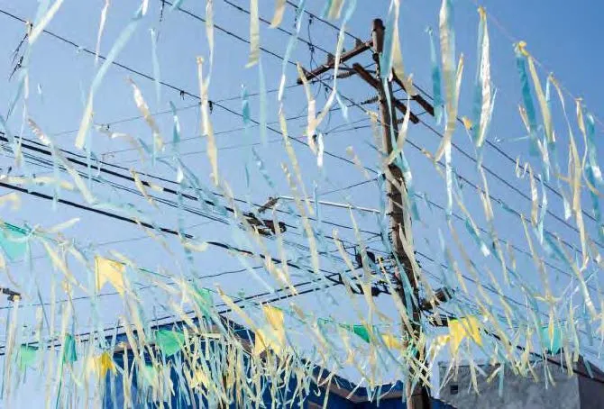 Fios coloridos azul, amarelo e branco pendurados com bandeiras triangulares e fitas finas em postes elétricos sob céu azul.