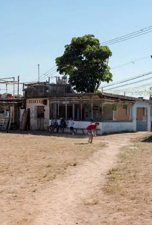 Crianças brincando em área de terra seca próxima a casa simples com grandes árvores ao fundo e céu claro.