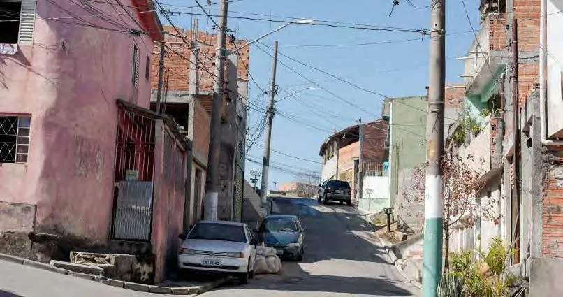 Rua inclinada em área urbana com casas simples, carros estacionados e muitos fios elétricos visíveis acima.
