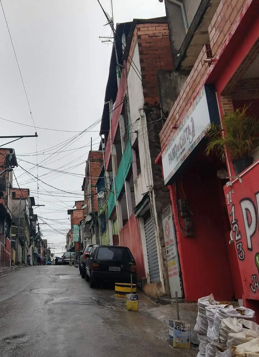 Rua inclinada com prédios residenciais simples, carros estacionados e fiação elétrica exposta em área urbana.