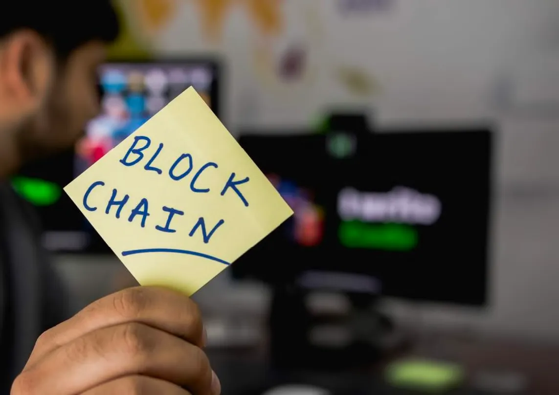 Pessoa segurando um post-it amarelo com a palavra 'BLOCKCHAIN' escrita à mão em azul, com monitores de computador desfocados ao fundo.