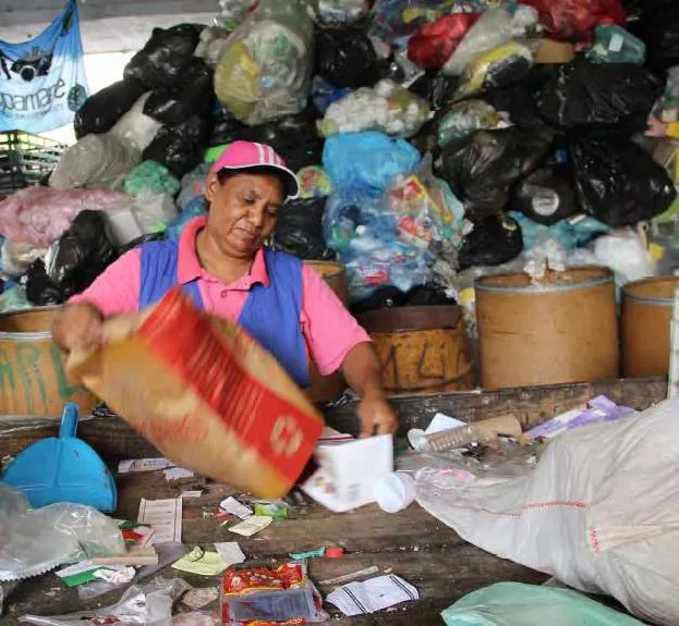 Mulher trabalhando na separação de resíduos recicláveis em um centro de coleta, com sacos de lixo empilhados ao fundo.