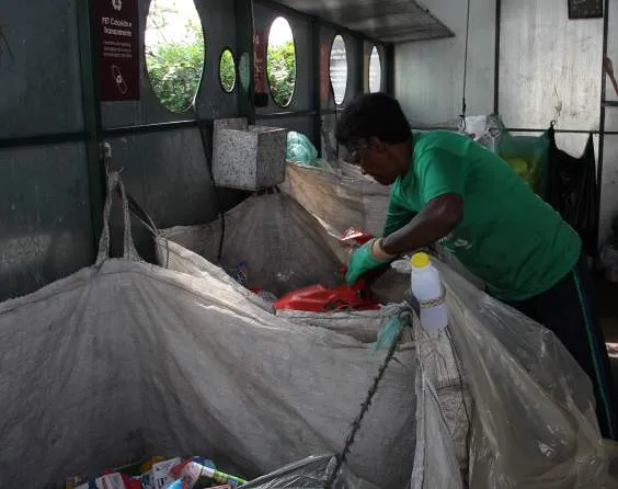 Homem com luvas verdes separando resíduos de recicláveis em grande recipiente dentro de instalação de coleta seletiva.