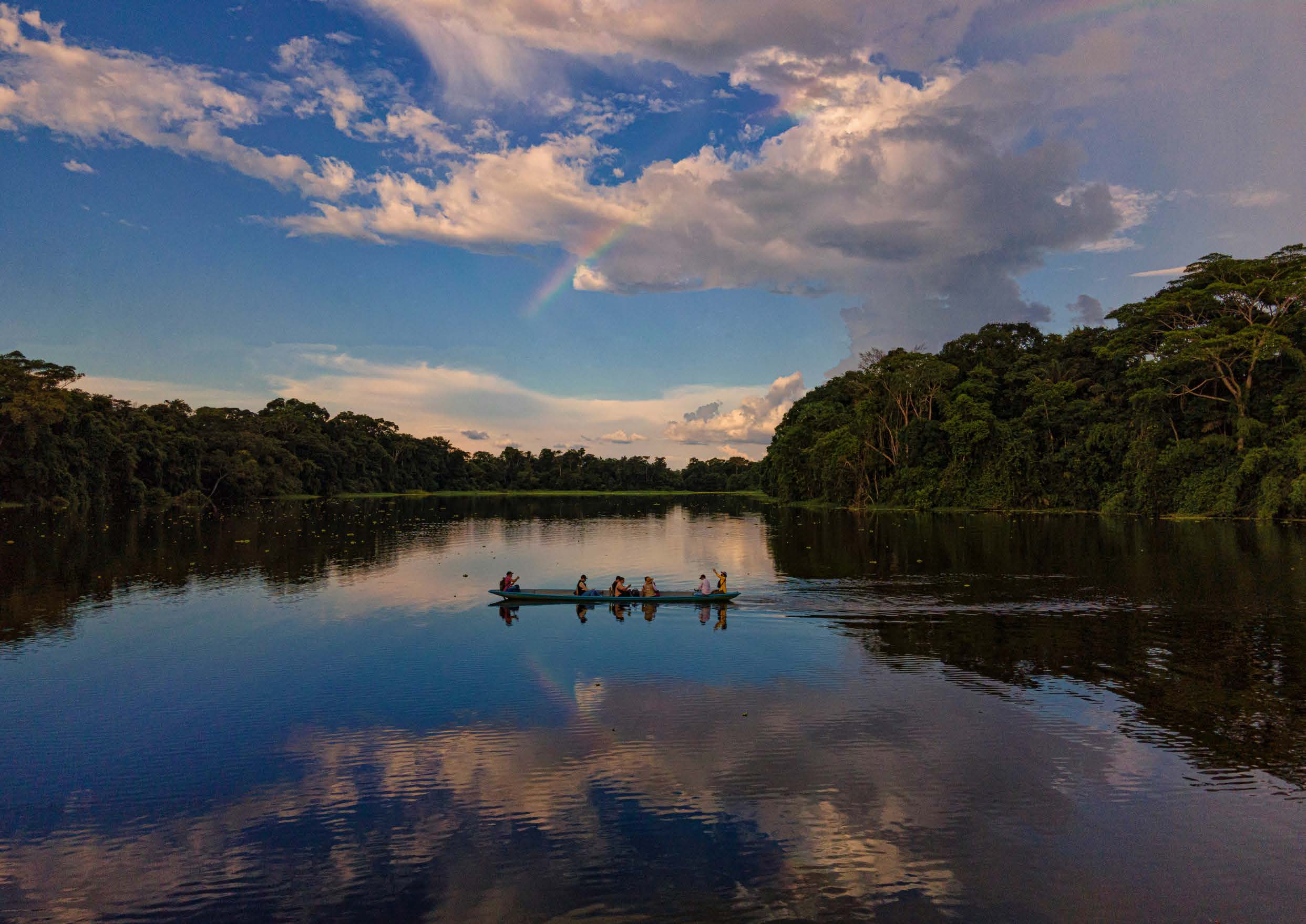 Seis pessoas remando em uma canoa no rio calmamente refletindo o céu azul com nuvens e um arco-íris, cercado por densa floresta verde.