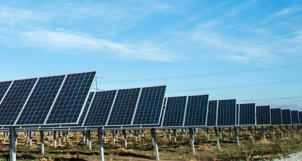 Painéis solares alinhados em um campo sob céu azul com poucas nuvens.
