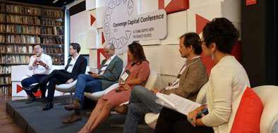 Mesa redonda com cinco pessoas discutindo em um evento do Cambridge Capital Conference, com estantes de livros ao fundo.